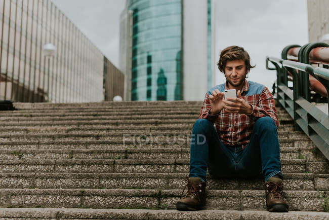 Porträt eines lächelnden bärtigen Mannes in lässiger Kleidung, der auf den Stufen der Straße sitzt und das Telefon benutzt — Stockfoto