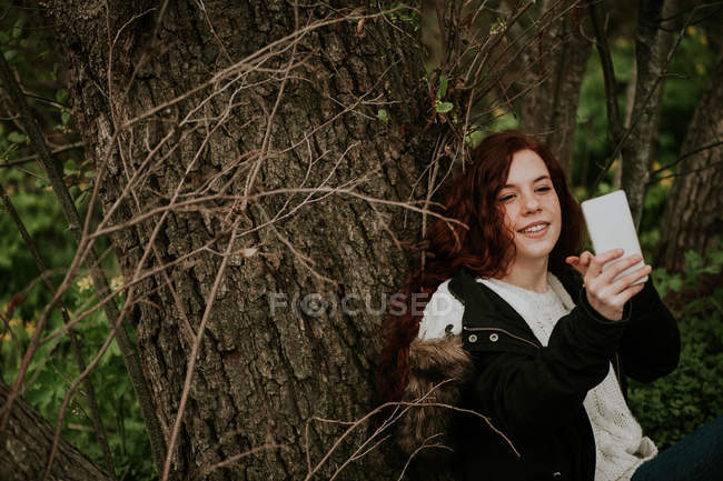 Улыбающаяся девушка делает селфи за деревом в лесу — стоковое фото