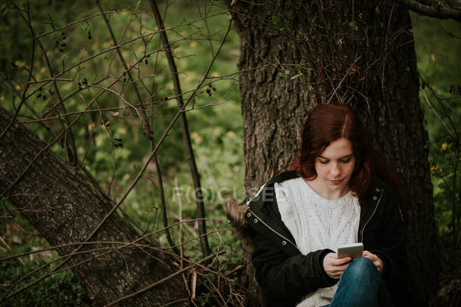 Рыжая девушка сидит у дерева и просматривает смартфон — стоковое фото