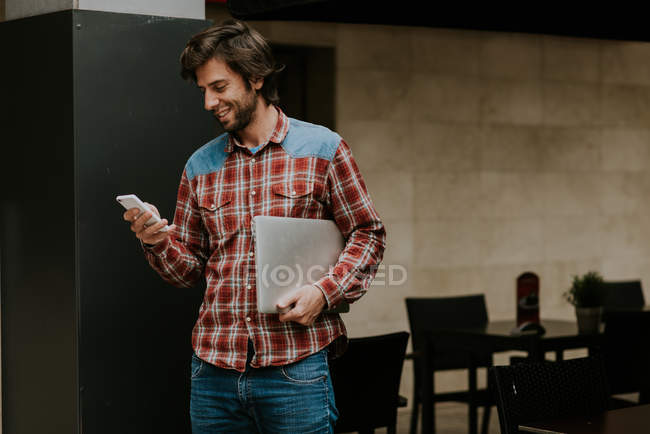 Side view of brunet man in checkered shirt carrying laptop and using smartphone — Stock Photo
