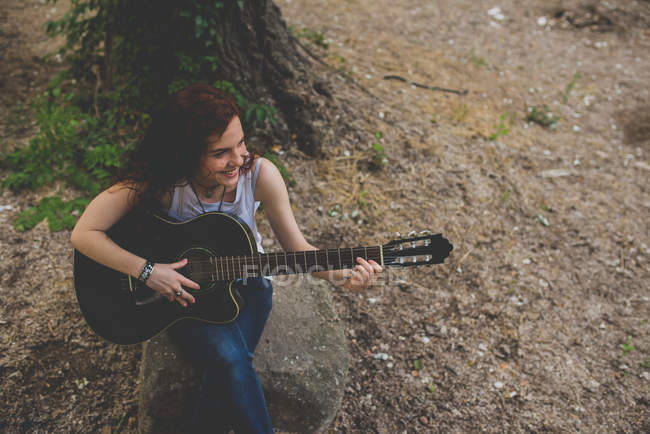 Portrait grand angle de fille ridée souriante assise sur le rock et jouant de la guitare — Photo de stock