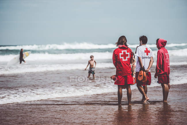Задний вид трех спасателей в красной форме, стоящих на пляже — стоковое фото