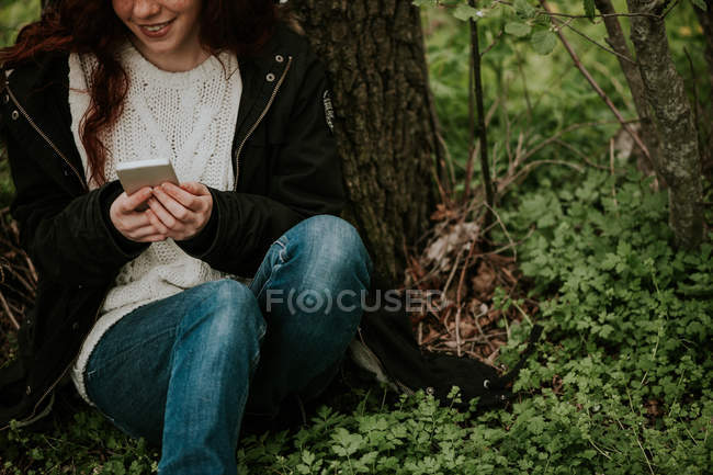 Cultivo chica jengibre sentado junto al árbol y el teléfono inteligente de navegación - foto de stock