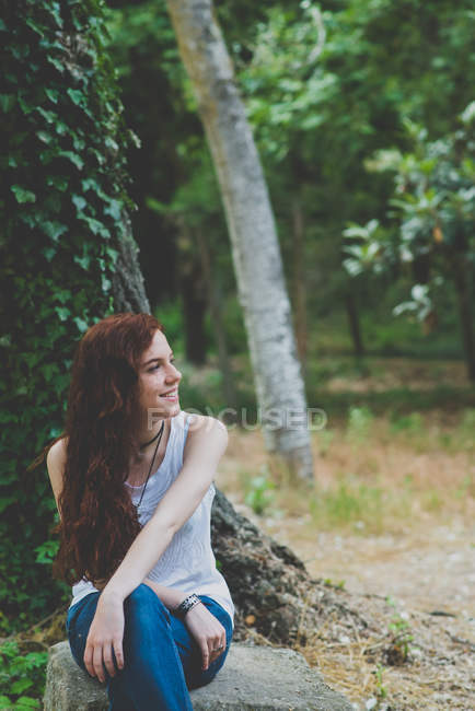 Portrait of smiling freckled girl sitting on stone and looking aside at countryside woods — Stock Photo