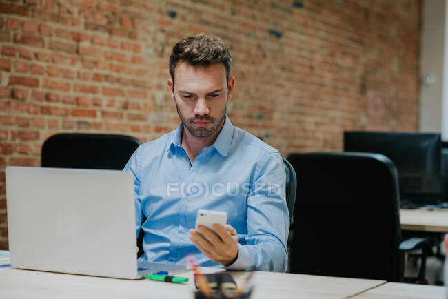 Homme utilisant son smartphone alors qu'il était assis à son ordinateur portable au bureau. Plan intérieur horizontal — Photo de stock