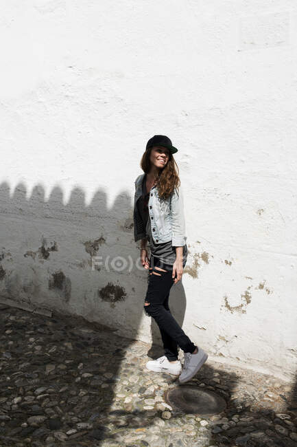 In voller Länge Aufnahme der jungen stilvollen Frau lächelnd und posierend vor schäbiger weißer Wand. — Stockfoto