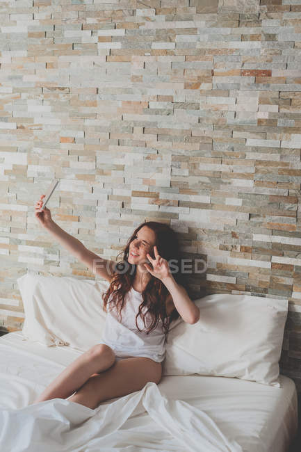 Chica sentada en la cama y tomando selfie - foto de stock