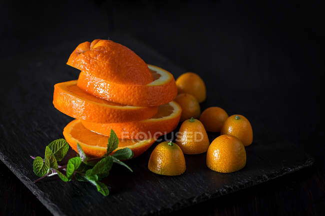 Naranja fresca en rodajas y kumquats a la mitad en pizarra con hojas de menta - foto de stock
