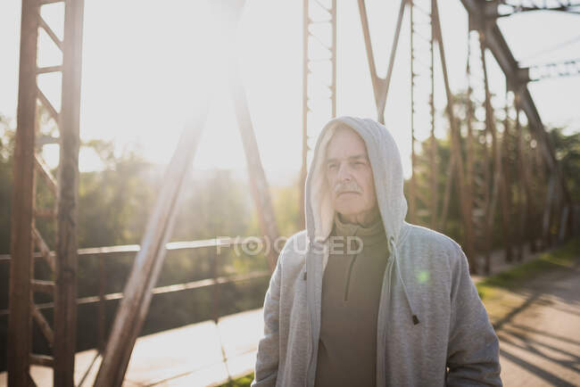 Vista del anciano en sudadera con capucha parado en el puente en un día soleado. Espacio de copia - foto de stock