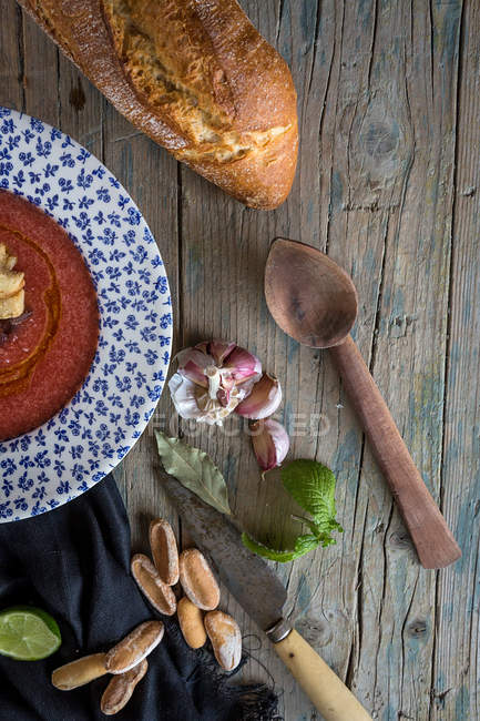 Soupe aux tomates dans une assiette à motifs sur une table en bois avec du pain et des ingrédients — Photo de stock
