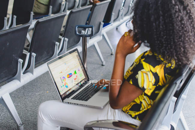 Side view of woman sitting at waiting room and using laptop on knees and talking on smartphone — Stock Photo