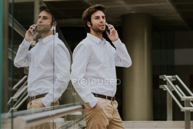Glass fence reflecting brunette businessman in white shirt talking on smartphone at urban scene — Stock Photo