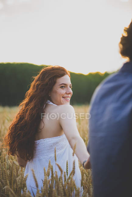 Вид сзади на девушку с вьющимися рыжими волосами, идущую по ржаному полю и оглядывающуюся через плечо на парня — стоковое фото
