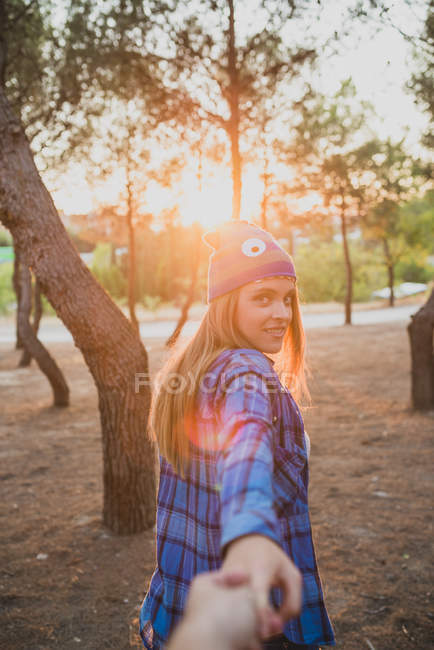 Retrato de menina em chapéu de malha engraçado olhando sobre o ombro e guiando com puxar a mão na luz do pôr do sol na floresta — Fotografia de Stock