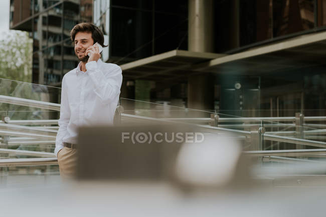 Портрет улыбающегося бизнесмена в белой рубашке, идущего рядом с бизнес-зданием и разговаривающего по смартфону — стоковое фото