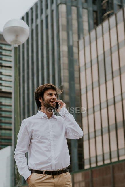 Low angle portrait of smiling businessman in white shirt talking on smartphone over business building facade at backdrop — Stock Photo