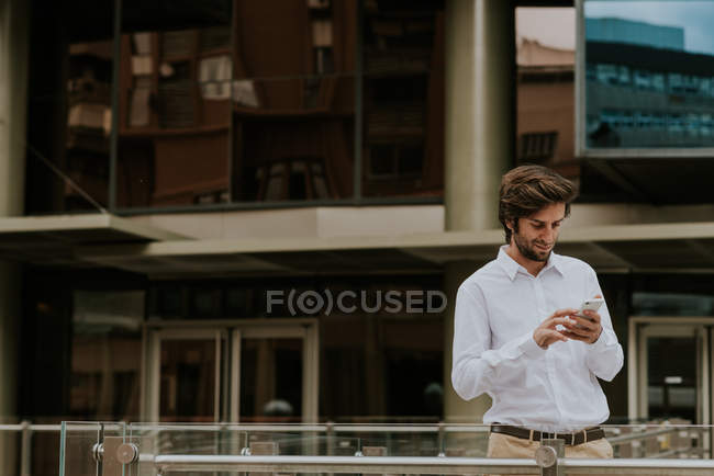 Portrait of smiling businessman in white shirt using phone over business building glass facade on background — Stock Photo