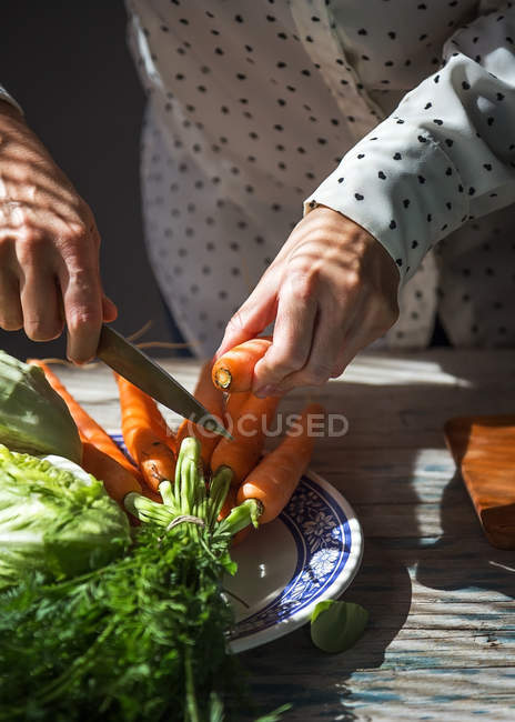 Close up view of female hands cutting of with knife carrots leaves over plate on kitchen table — Stock Photo