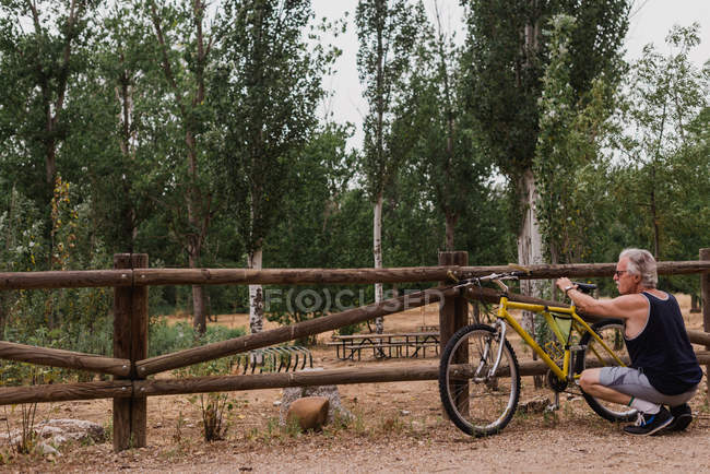 Vista trasera del hombre mayor en cuclillas cerca apoyado en la bicicleta de la cerca rural y ajuste sentarse - foto de stock
