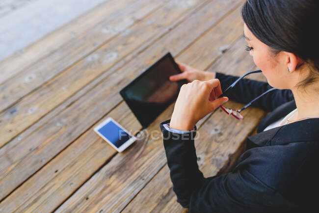 Rear view of woman sitting at wooden table and using tablet while biting earpiece of glasses. Copyspace — Stock Photo