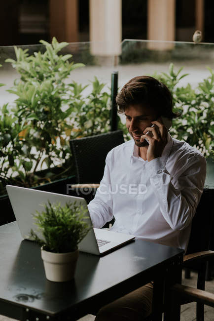 Retrato de un hombre de negocios sonriente sentado en la mesa y hablando por teléfono inteligente mientras usa el portátil en la terraza de la cafetería - foto de stock