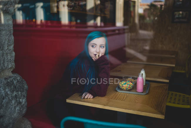 Portrait of girl with blue hair sitting at table with yogurt and bowl of cereals on tray and looking at window — Stock Photo