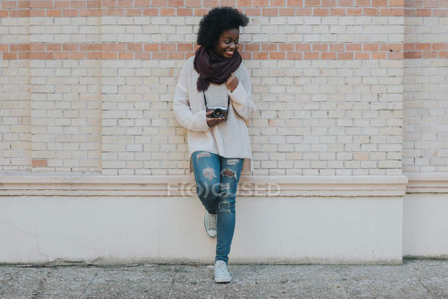 Portrait of smiling girl with camera hanging on neck leaning on brick wall and looking away — Stock Photo