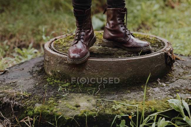 Imagem cortada de pernas masculinas em botas elegantes em pé no musgo esgoto coberto bueiro — Fotografia de Stock