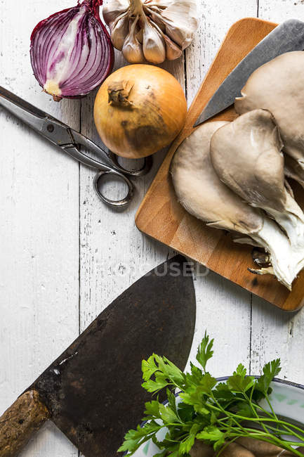 Top view of board with pleurotus mushrooms and chopper on white rustic table with onion and garlic — Stock Photo