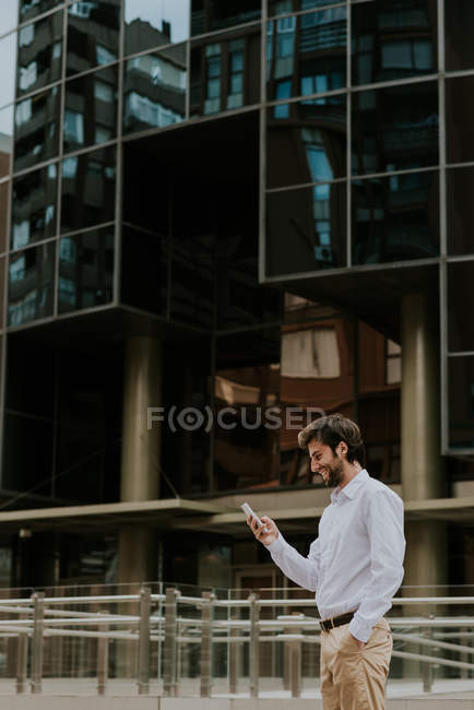 Vista lateral de un hombre de negocios sonriente con camisa blanca usando un teléfono inteligente cerca de la fachada del edificio moderno - foto de stock