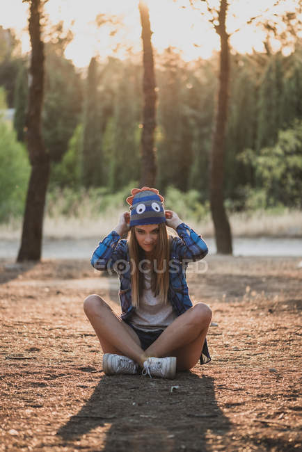 Retrato de mulher sentada no chão na floresta e ajustando chapéu de lã engraçado — Fotografia de Stock