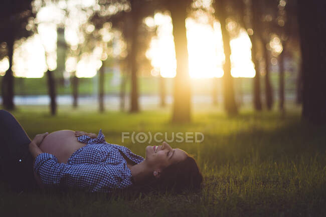Happy pregnant woman lying on grass in the park. Horizontal outdoors shot. — Stock Photo