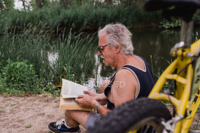 Side view of senior man sitting on forest river shore and reading book beside bicycle — Stock Photo