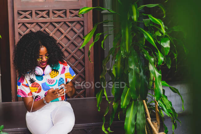Retrato de una chica con una camiseta colorida sentada cerca de una planta en maceta y charlando en un teléfono inteligente - foto de stock