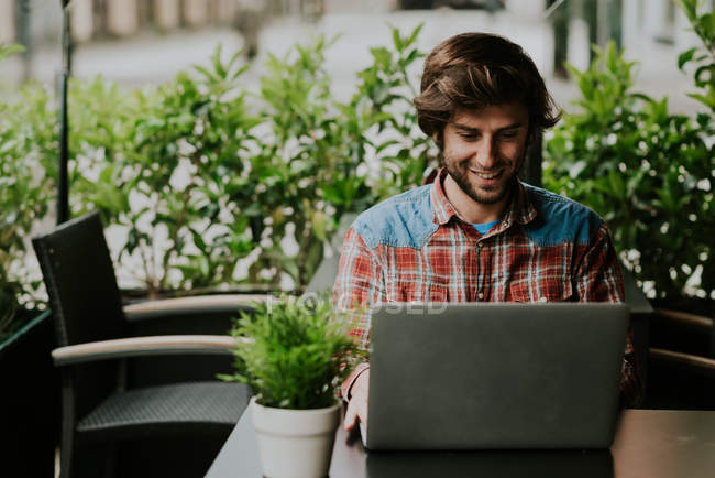 Portrait of bearded man sitting at cafe terrace table with potted plant and using laptop — Stock Photo