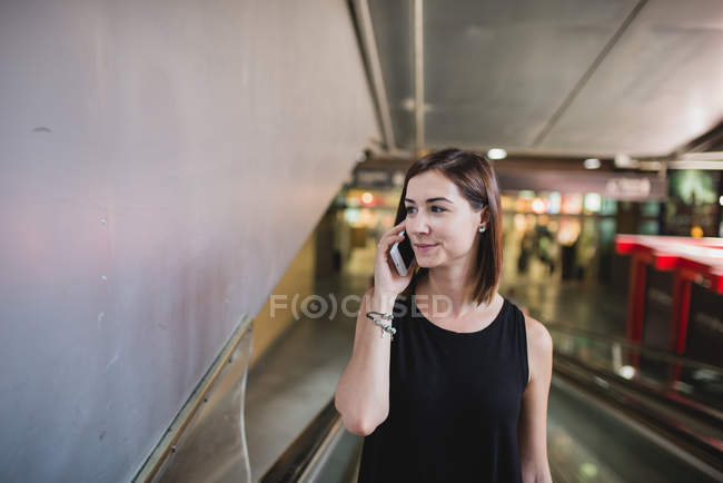 Retrato de mujer joven moviéndose en escaleras mecánicas y hablando en el teléfono inteligente en la estación de tren - foto de stock