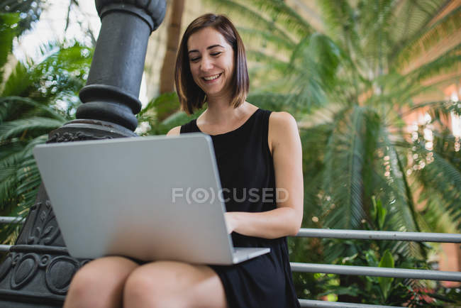 Низкоугольный портрет улыбающейся девушки, сидящей и использующей ноутбук на коленях — стоковое фото