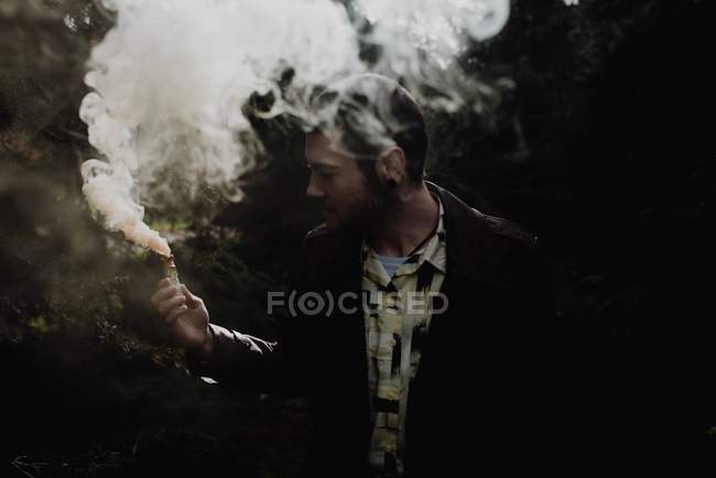 Портрет молодого человека, стоящего в лесу и смотрящего на дымовую свечу в руке — стоковое фото