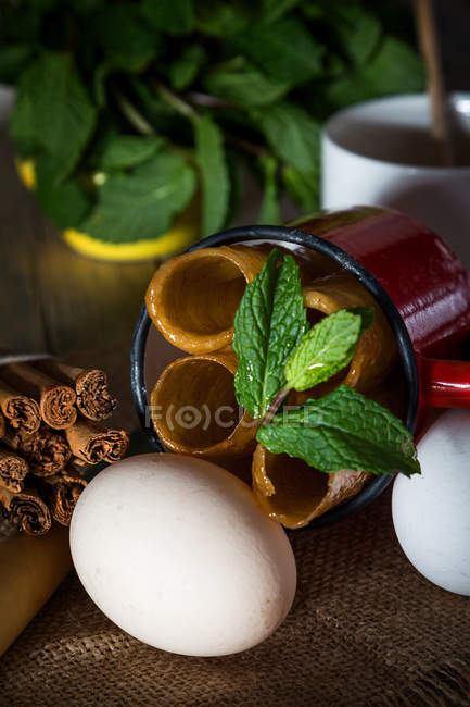 Still life of fried honey dough tubes with mint in mug lying on sacking with eggs — Stock Photo