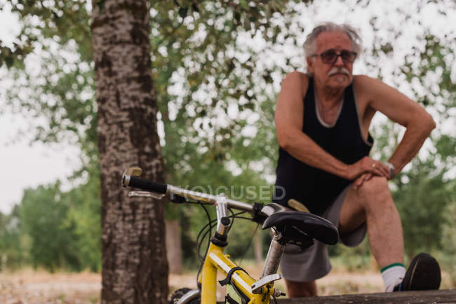 Vista frontal del anciano estirando las piernas en el parque cerca de la bicicleta - foto de stock