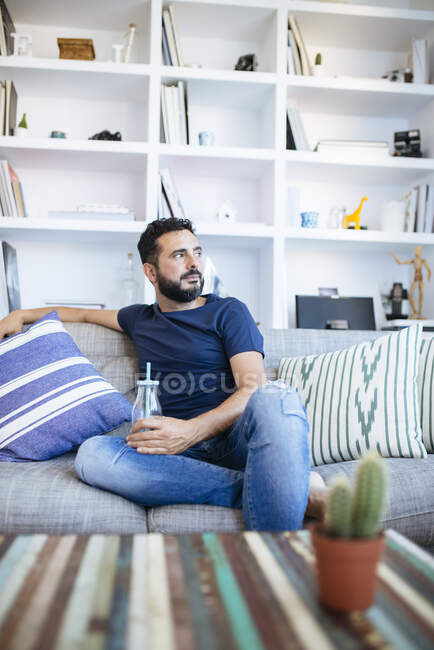Brunet sitting on sofa with bottle with straw in his arm and looking away. — Stock Photo