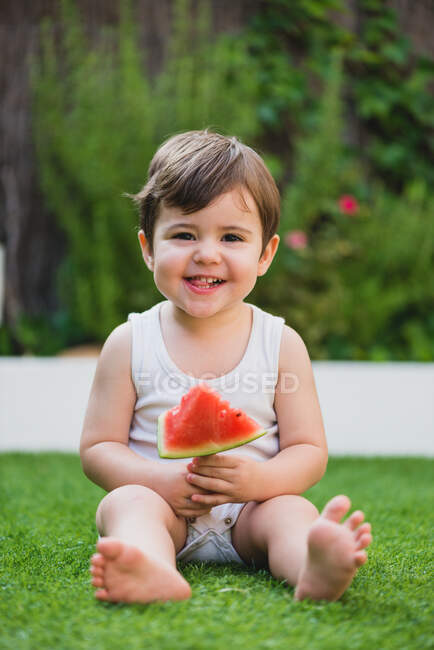 Retrato de adorable bebé sonriendo felizmente a la cámara con rebanada de deliciosa y jugosa sandía. - foto de stock