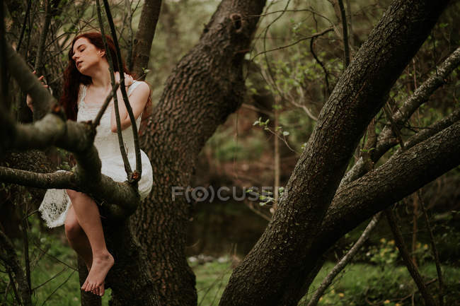 Ginger girl in white dress sitting on branches and touching neck — Stock Photo