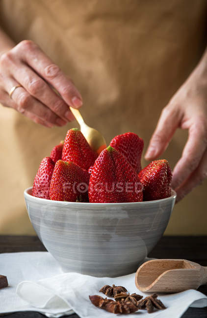 Imagen de la cosecha de manos femeninas poniendo fresas en un tazón en la mesa con estrellas de anís y cuchara - foto de stock