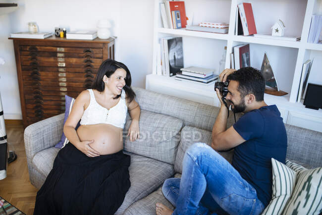 Adult husband taking a camera shot of his wife, sitting on sofa in living room. — Stock Photo