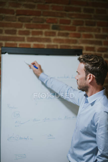 Un hombre serio escribiendo en una pizarra en la oficina. Tiro vertical en interiores - foto de stock