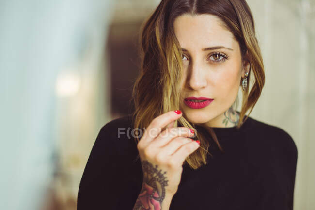 Glamour woman with a tattooed arm holding hair and looking at camera. Horizontal indoors shot. — Stock Photo