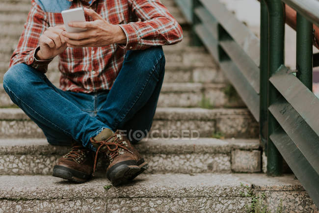 Cropped image of male in checked shirt sitting on steps and using smartphone — Stock Photo