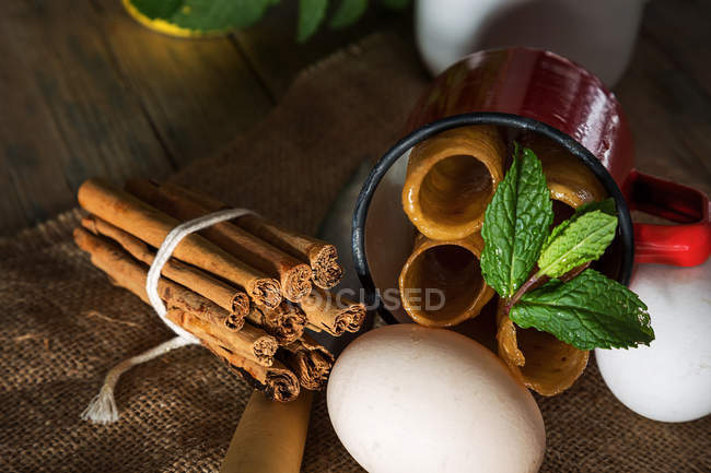 Still life of fried honey dough tubes with mint in mug lying on sacking with eggs and cinnamon sticks — Stock Photo