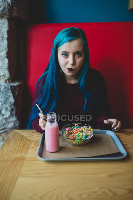 Retrato de adolescente de pelo azul sentada en la mesa de la cafetería con yogur y tazón de cereales de colores en la bandeja y mirando a la cámara - foto de stock